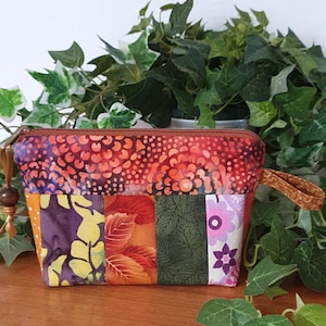 Könnte beinhalten: Eine bunte Patchwork-Kosmetiktasche mit braunem Reißverschluss und Handschlaufe. Die Tasche besteht aus verschiedenen gemusterten Stoffen in Orange, Lila, Grün und Rosa. Der obere Bereich hat ein Blumenmuster.