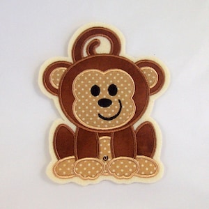 Könnte beinhalten: Ein braunes Filz-Affe-Applikationsmotiv mit weißem Punktmuster. Der Affe sitzt mit seinem Schwanz über dem Kopf und hat ein lächelndes Gesicht.