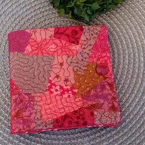 Könnte beinhalten: Eine Patchwork-Stoffbrieftasche in Rosa-, Rot- und Brauntönen. Die Brieftasche besteht aus verschiedenen Stoffstücken, die mit dekorativen Nähten zusammengenäht sind. Ein großer, runder, rosa Knopf verschließt die Brieftasche. Das Gesamtdesign ist eine Mischung aus floralen und abstrakten Mustern.