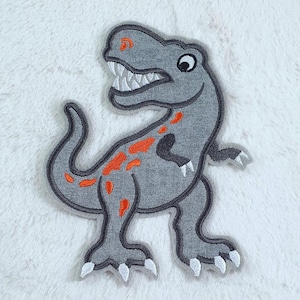 Könnte beinhalten: Ein gestickter Dinosaurier-Patch in Grau mit orangefarbenen Akzenten. Der Dinosaurier steht auf allen Vieren, mit offenem Maul und sichtbaren Zähnen.