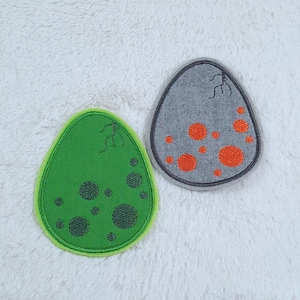 Könnte beinhalten: Zwei gestickte Filzpatches in Form von Eiern. Einer ist grün mit dunkelgrünen Punkten und der andere ist grau mit orangefarbenen Punkten.