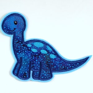 Könnte beinhalten: Ein blauer Filz-Dinosaurier-Aufnäher auf weißem Hintergrund. Der Dinosaurier hat ein gepunktetes Muster und ein freundliches Gesicht.