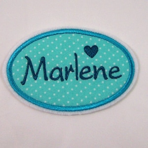Könnte beinhalten: Ein ovaler Patch in Türkisblau mit weißen Punkten und einem dunkelblauen gestickten Herzen. Der Name "Marlene" ist in dunkelblauer Kursivschrift gestickt.