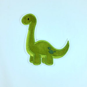Könnte beinhalten: Grünfilzene Dinosaurier-Applikation mit weißem Umriss. Der Dinosaurier hat einen langen Hals und einen kleinen Kopf mit einem schwarzen Auge.