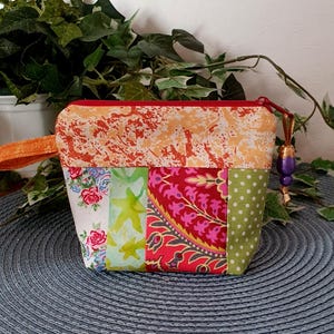 Könnte beinhalten: Ein Patchwork-Reißverschlusstasche mit verschiedenen Stoffmustern. Der obere Teil ist orange mit einem gesprenkelten Design, während der Körper Blumen-, Stern- und Tupfenmuster in Rot, Grün und Pink aufweist. Sie hat einen kleinen orangefarbenen Griff und einen lila Perlen-Reißverschluss.