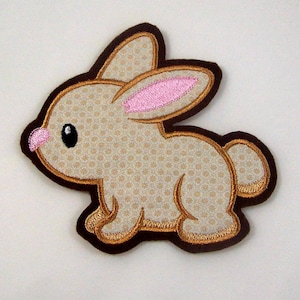Könnte beinhalten: Ein gestickter Hasen-Patch in Braun und Beige mit rosa Ohren. Der Hase blickt nach rechts und hat einen braunen Umriss.