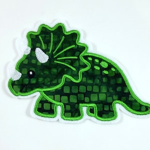 Könnte beinhalten: Ein grüner Triceratops-Aufnäher mit weißem Rand. Der Dinosaurier hat einen strukturierten, quadratisch gemusterten Körper und weiße Hörner. Das Design hat eine detaillierte, gestickte Kontur.