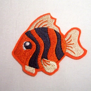 Könnte beinhalten: Gestickter Aufnäher eines Cartoon-Fisches in Orange, Marineblau und Beige. Der Fisch hat ein großes Auge und ist orange umrandet. Der Körper des Fisches weist marineblaue Streifen und beige Flossen auf.