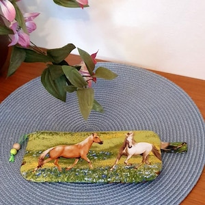 Könnte beinhalten: Ein rechteckiges Stoffetui mit Reißverschluss, bedruckt mit einer Szene, die ein hellbraunes und ein weißes Pferd zeigt, die durch eine grüne Wiese mit gelben und blauen Wildblumen laufen. Am Reißverschluss befindet sich eine grüne Kordel mit Holzperlen.