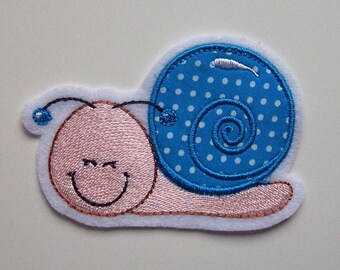 Könnte beinhalten: Eine gestickte Schnecken-Applikation in Rosa und Blau auf weißem Hintergrund. Die Schnecke hat eine blaue Schale mit weißen Punkten und einen rosa Körper mit einem lächelnden Gesicht.