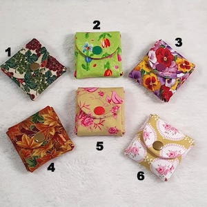 Könnte beinhalten: Sechs kleine, quadratische Stoffbeutel mit Knopfverschlüssen werden gezeigt. Die Beutel weisen verschiedene Blumen- und Blättermuster in Farben wie Rot, Grün, Gelb und Orange auf. Jeder Beutel ist etwa 7,6 cm groß.