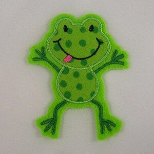 Könnte beinhalten: Grünfilz-Frosch-Applikation mit einem lächelnden Gesicht und einer herausgestreckten rosa Zunge. Der Frosch hat grüne Punkte und ist mit grünem Garn umrandet.