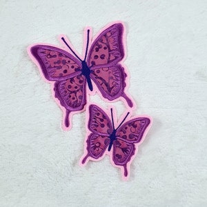 Könnte beinhalten: Zwei lilafarbene Bügelbilder in Form von Schmetterlingen mit rosa Umrandungen. Der größere Schmetterling hat ein detaillierteres Design mit einem Muster aus kleinen Kreisen.