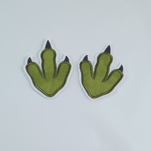 May include: Two green dinosaur footprints with black claws. The footprints are embroidered and have a white outline.