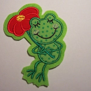 Könnte beinhalten: Grüne Filzapplikation eines Frosches, der eine rote Blume hält. Der Frosch hat ein lächelndes Gesicht und geschlossene Augen. Die Blume hat eine gelbe Mitte.