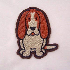 Könnte beinhalten: Gestickter Aufnäher mit einem Cartoon-Beagle-Hund. Der Aufnäher hat einen braunen Rand, der Körper des Hundes ist beige, die Ohren braun und das Gesicht weiß und braun. Der Hund sitzt mit eingerollter Rute.