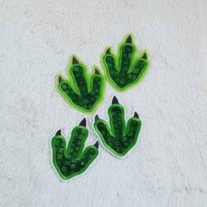 Könnte beinhalten: Vier grüne Dinosaurier-Fußabdrücke mit schwarzen Krallen. Die Fußabdrücke sind aus Stoff und haben ein strukturiertes Design.