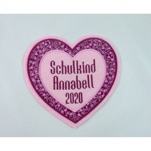 Könnte beinhalten: Rosa herzförmiger Patch mit burgunderfarbener Blumenbordüre. Der Text "Schulkind Annabell 2020" ist in Weiß auf den Patch gestickt.