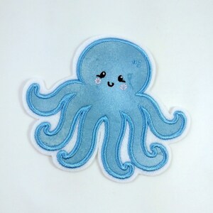 Könnte beinhalten: Ein hellblauer gestickter Oktopus mit einem lächelnden Gesicht und glitzernden Augen. Der Oktopus hat acht Tentakel.