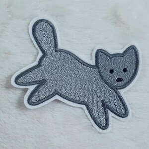 Könnte beinhalten: Ein grauer Filz-Katzen-Applikationen mit einem einfachen Design. Die Katze blickt nach rechts und hat einen kleinen, runden Kopf mit zwei kleinen, schwarzen Augen und einer schwarzen Nase.