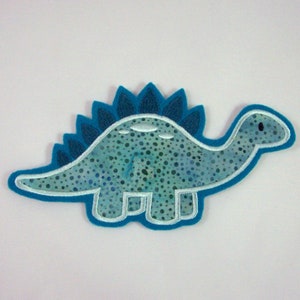 Könnte beinhalten: Eine blaue Filz-Dinosaurier-Applikation mit weißem Umriss und einem gepunkteten Muster. Der Dinosaurier hat einen stacheligen Rücken und ein freundliches Gesicht.