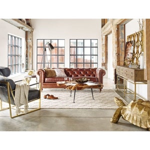 May include: A brown leather tufted sofa with a wooden frame and gold legs sits in a living room with a large, rustic wooden beam overhead. A gold metal coffee table with a live edge top sits in front of the sofa. A gold metal chair with a black cushion sits to the left of the sofa. A gold metal console table with drawers sits to the right of the sofa. A gold metal turtle sculpture sits on the floor in front of the console table.