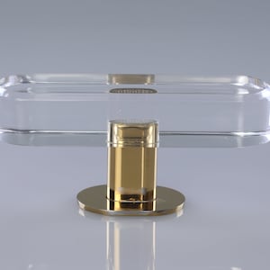 May include: A clear acrylic rectangular tray with rounded edges sits atop a gold metal base. The base has a cylindrical shape with a round top and a flat bottom.