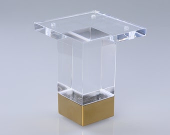 Könnte beinhalten: Klares Acryl-Möbelbein mit goldfarbener Metallbasis. Das Bein ist quadratisch und hat eine flache Oberseite.
