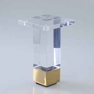 Könnte beinhalten: Klares Acryl-Möbelbein mit goldfarbener Metallbasis. Das Bein ist quadratisch und hat eine flache Oberseite.