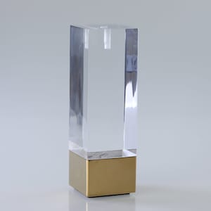 Op de afbeelding: Een transparant acryl meubelleg met een gouden metalen basis. Het been is vierkant en heeft een schroefdraadgat aan de bovenkant.