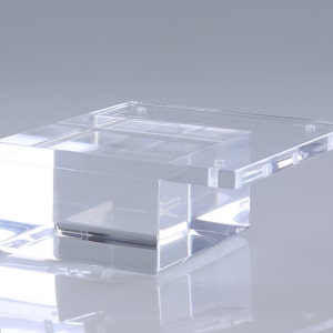 Peut inclure: Un présentoir en acrylique transparent avec une base carrée et un dessus rectangulaire. Le dessus est légèrement décalé de la base.