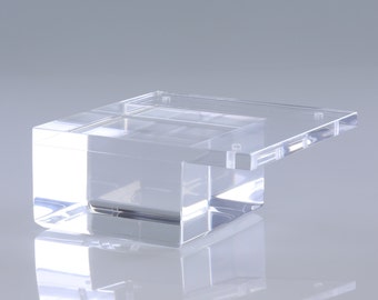 Könnte beinhalten: Ein durchsichtiger Acryl-Präsentationsständer mit quadratischer Basis und rechteckiger Oberseite. Die Oberseite ist leicht versetzt zur Basis.