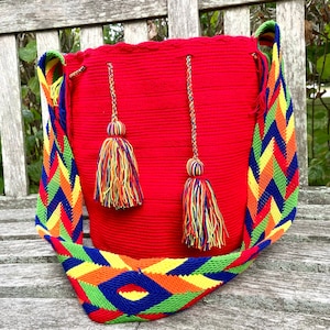 May include: A red crocheted bag with a colorful woven strap and tassels. The bag is sitting on a wooden bench.