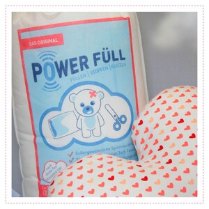 May include: A white bag with a blue and red label that says "DAS ORIGINAL POWER F&Uuml;LL" and "F&Uuml;LLEN STOPFEN BASTELN". The bag is next to a white pillow with a red heart pattern.