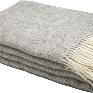 Peut inclure: Une couverture en laine gris chiné à motif chevron avec une frange blanche. La couverture est pliée et a une apparence douce et texturée.