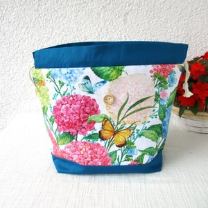 Könnte beinhalten: Eine türkisfarbene Kordeltasche mit weißem Hintergrund und einem Blumendruck mit rosa, weißen und blauen Hortensien und Schmetterlingen. Die Tasche hat einen Verschluss aus Holz.