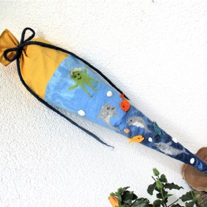 Könnte beinhalten: Ein blauer Filz-Kegel mit gelber Stoff-Oberseite und einem Kordelzug. Der Kegel ist mit Filz-Meerestieren verziert, darunter ein Oktopus, Delfine und Fische. Außerdem befinden sich weiße Filzpunkte auf dem Kegel.