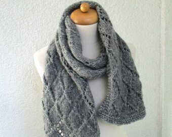 May include: A gray knitted scarf with a diamond pattern is draped over a mannequin. The scarf is made of a soft, textured yarn and has a delicate, openwork design along the edges. The scarf is approximately 70 inches long.