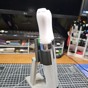 May include: A white and silver device with a black holder, supported by a gray stand. The device has a cylindrical body and a white top with two small openings. The holder is attached to a gray stand.