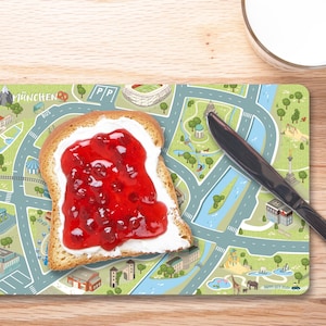 Può includere: Una fetta di pane tostato bianco con uno strato di formaggio fresco e marmellata di frutta rossa sopra. Il toast è su un sottopiatto con una mappa di una città stampata su di esso. Il sottopiatto è su una superficie di legno. Un coltello da burro argentato è sul tavolo.