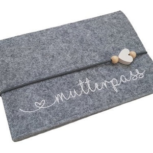 Könnte beinhalten: Graue Filztasche mit einem weißen gestickten Herzen und dem Text "Mutterpass". Die Tasche hat eine Holzperle und einen weißen Herzanhänger an einer schwarzen Schnur.