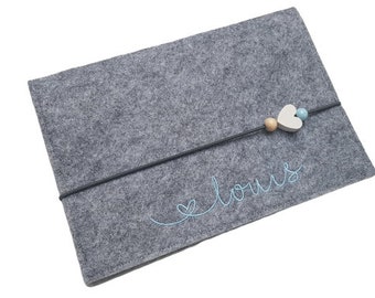 May include: A gray felt tablet sleeve with a black elastic band closure. The band is adorned with wooden beads and a heart-shaped charm. The name "louis" is embroidered in light blue on the front.