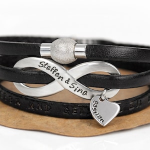 Könnte beinhalten: Ein schwarzes Lederarmband mit einem silberfarbenen Unendlichkeitssymbol, das mit dem Text Steffen & Sina graviert ist. Ein kleiner silberner Herzanhänger mit dem Namen Bastian hängt am Unendlichkeitssymbol, ergänzt durch eine strukturierte Silberperle.
