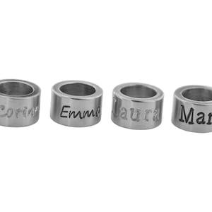 Könnte beinhalten: Vier zylindrische, silberfarbene Metallperlen in einer horizontalen Reihe. Jede Perle ist mit einem anderen Namen in schwarzer Schrift graviert: Corin, Emma, Laura und Marli. Die Perlen haben eine glatte, polierte Oberfläche und ein hohles Zentrum.