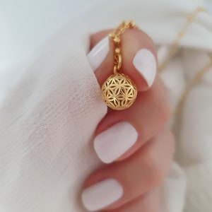 Könnte beinhalten: Eine goldene Halskette mit einem zarten, filigranen, runden Anhänger mit geometrischem Muster. Der Anhänger wird von einer Hand mit weißem Nagellack gehalten.