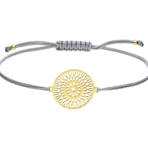 Puede incluir: Un brazalete ajustable de cuerda gris con un colgante circular dorado con un delicado dise&ntilde;o de corte intrincado.