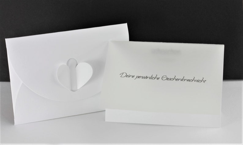 Puede incluir: Sobre blanco con un recorte en forma de coraz&oacute;n y una tarjeta blanca con el texto "Deine pers&ouml;nliche Geschenkernachricht" en negro.