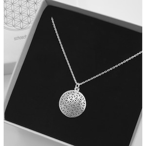 Könnte beinhalten: Eine silberne Halskette mit einem runden Anhänger, der ein detailliertes geometrisches Lebensblume-Design zeigt. Die Halskette wird in einer schwarzen Samtbox mit dem Wort "schosch" auf der Seite präsentiert.