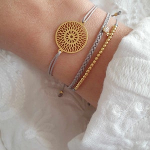 Puede incluir: Tres pulseras en una mu&ntilde;eca. La pulsera superior tiene un dije circular dorado con un dise&ntilde;o delicado e intrincado. La pulsera del medio es un cord&oacute;n trenzado gris. La pulsera inferior es una cadena dorada con peque&ntilde;as cuentas y un solo cristal de imitaci&oacute;n transparente.