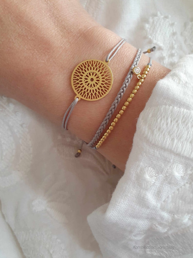 Puede incluir: Tres pulseras en una mu&ntilde;eca. La pulsera superior tiene un dije circular dorado con un dise&ntilde;o delicado e intrincado. La pulsera del medio es un cord&oacute;n trenzado gris. La pulsera inferior es una cadena dorada con peque&ntilde;as cuentas y un solo cristal de imitaci&oacute;n transparente.